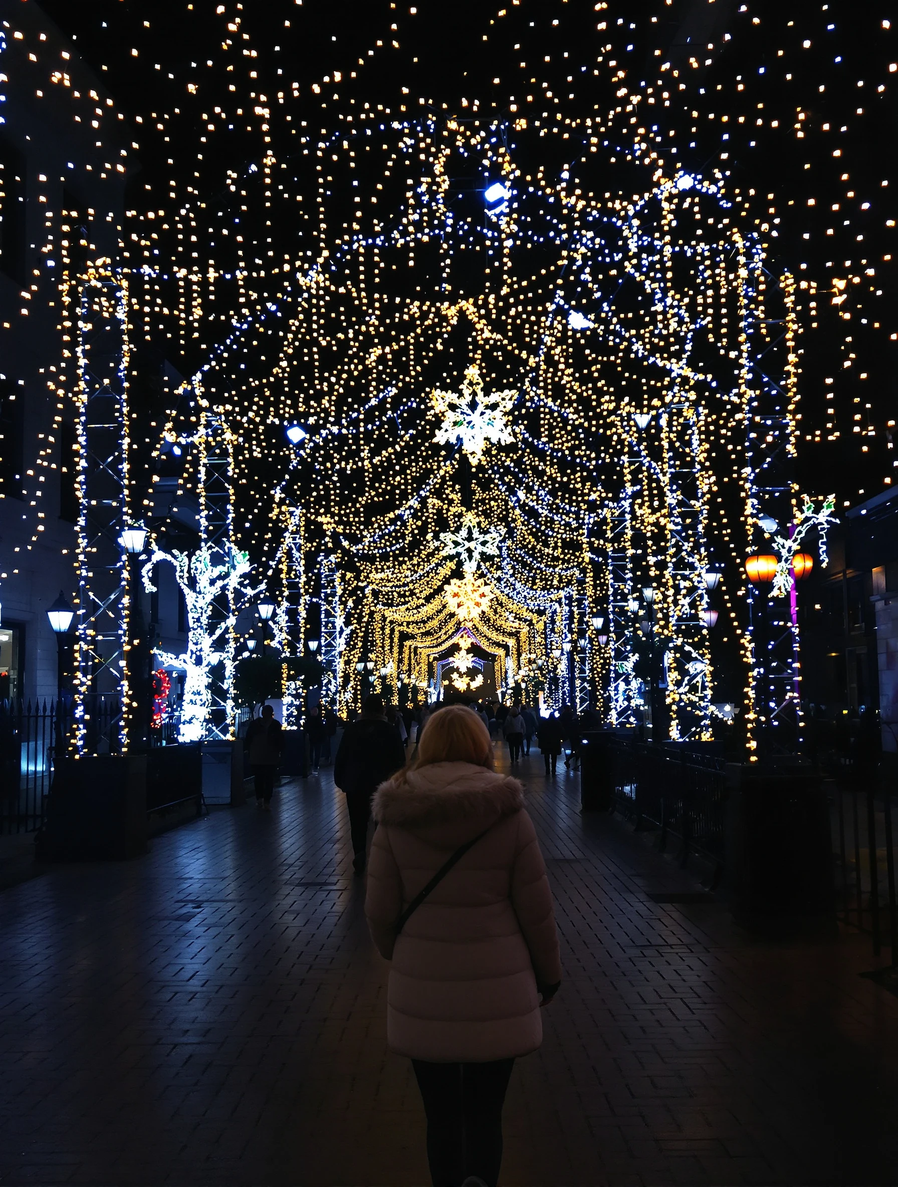 Woman walking through city decorated with Christmas lights at night. Winter coat, magical illumination, holiday spirit, urban winter wonderland.