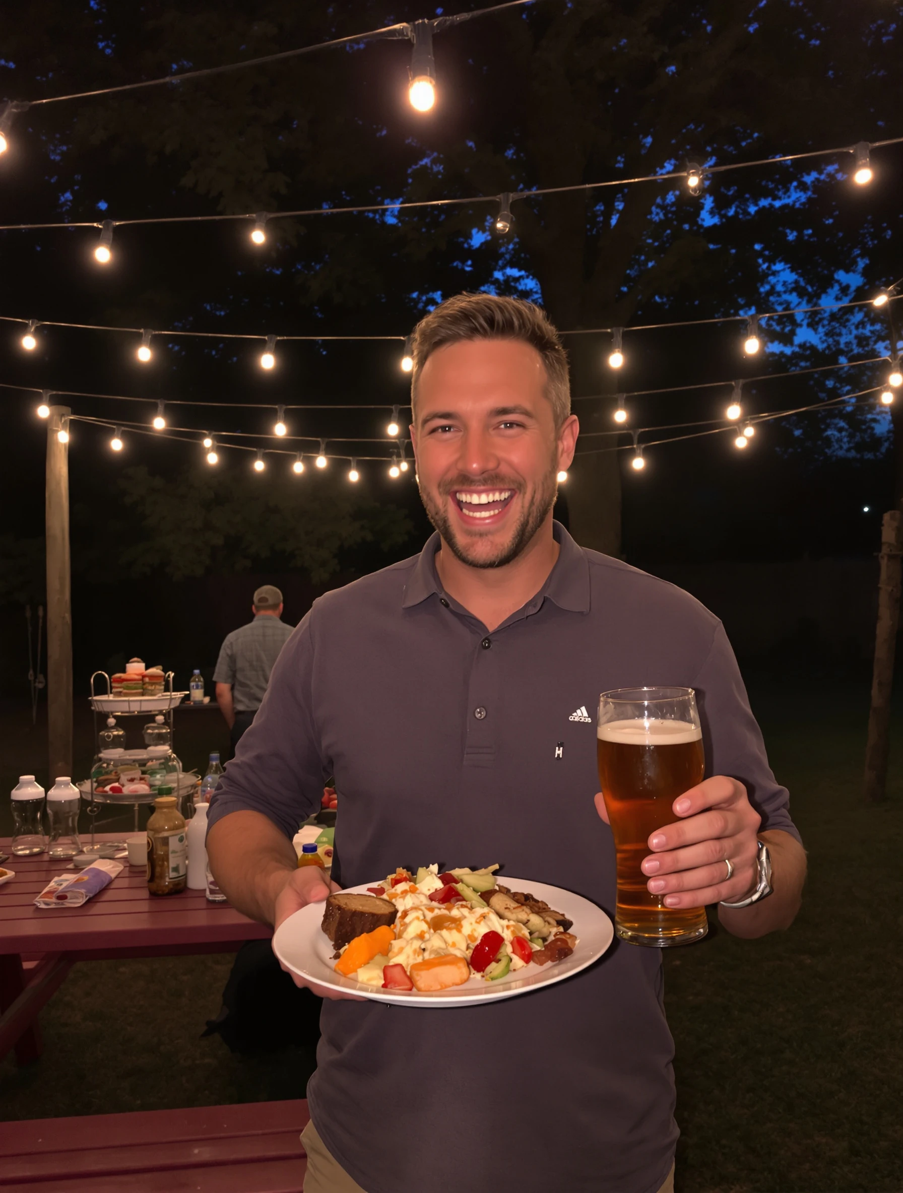 Man at a backyard barbecue birthday holding a plate of food and a beer, string lights overhead at dusk, big relaxed laugh, picnic table behind him.