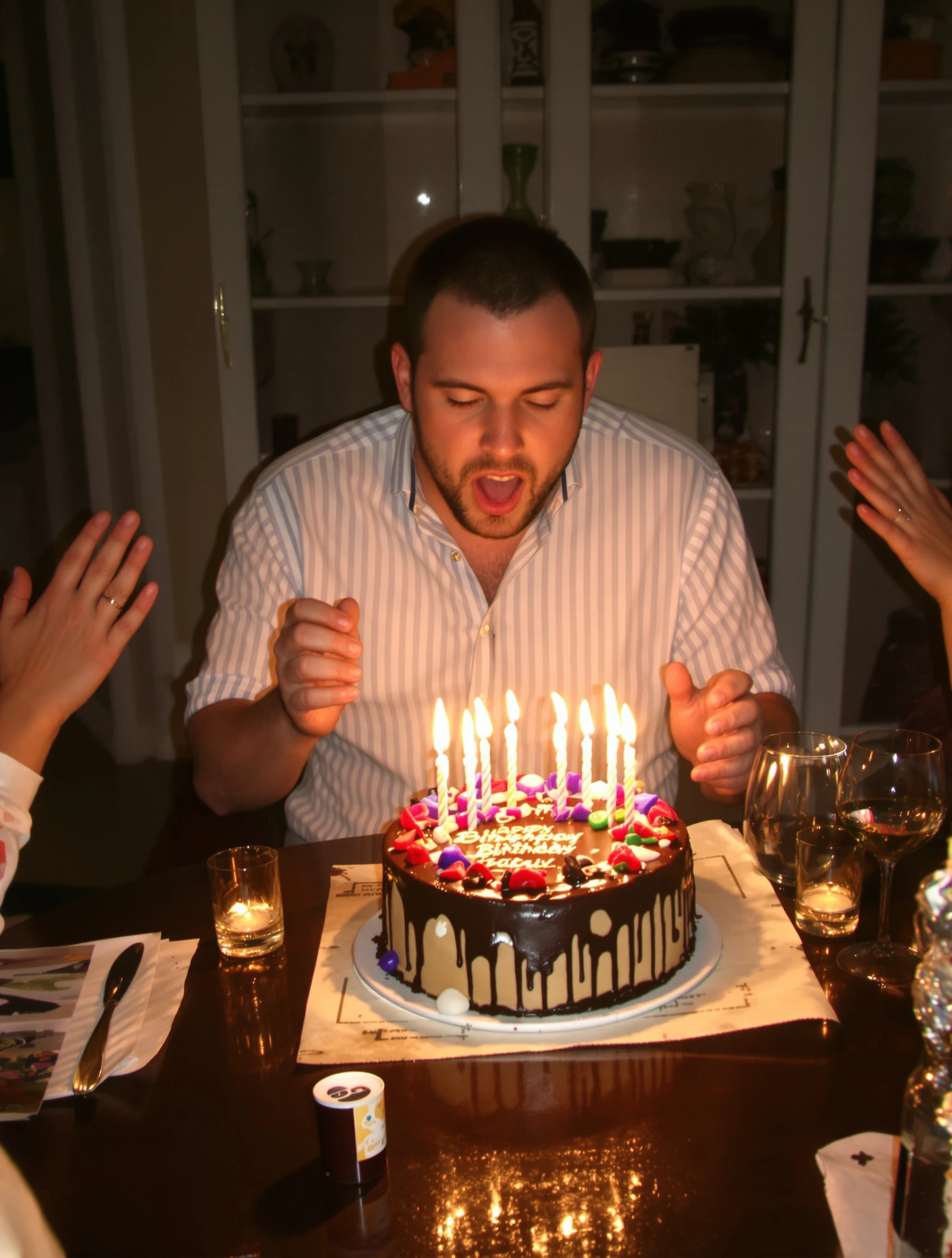 Man blowing out candles on a chocolate birthday cake at a cozy dinner table, warm candle glow on his face, friends' hands clapping at the edges of the frame.