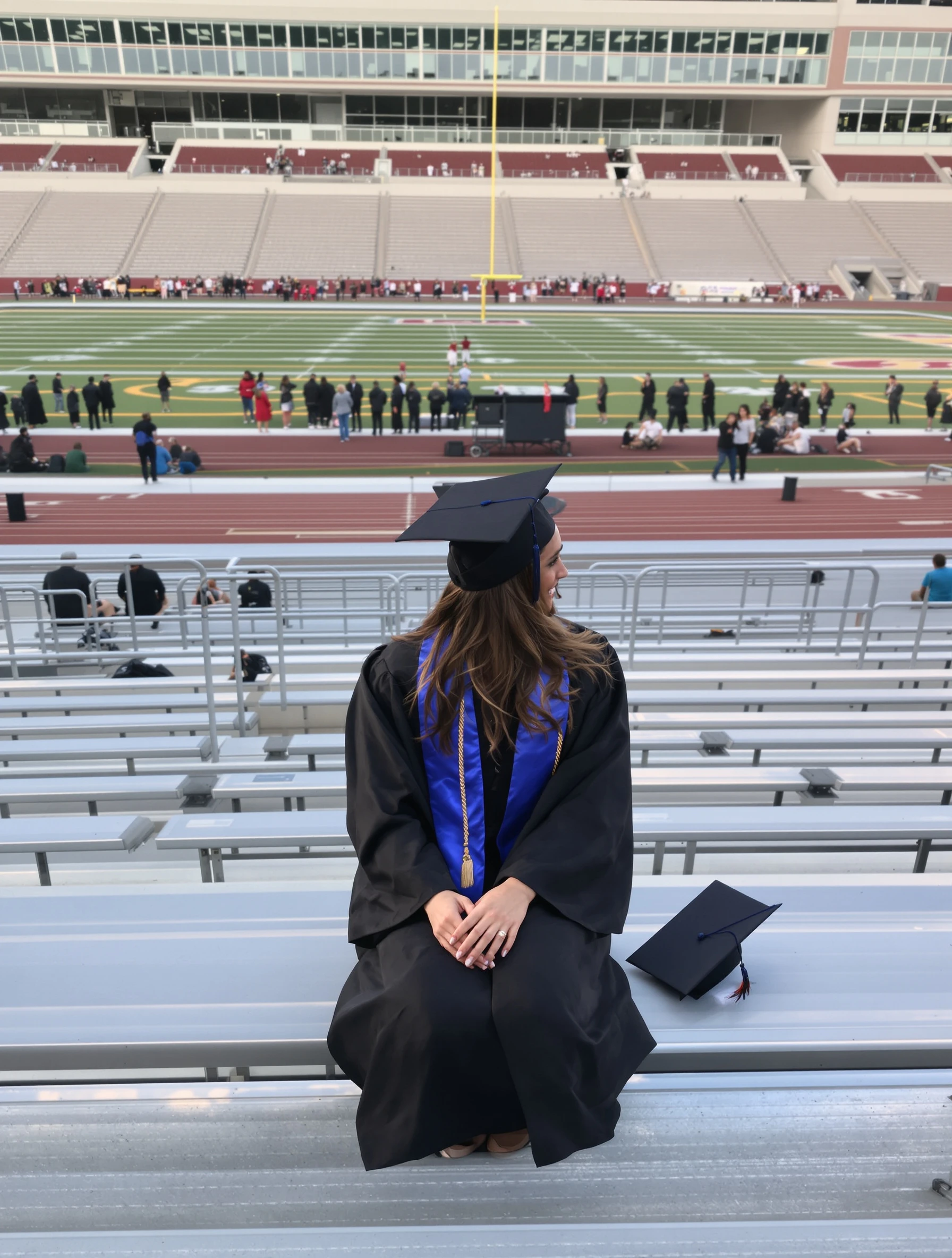 Woman in cap and gown seated on the bleachers of an empty football stadium, diploma beside her, looking out at the field with a reflective smile, late afternoon light.