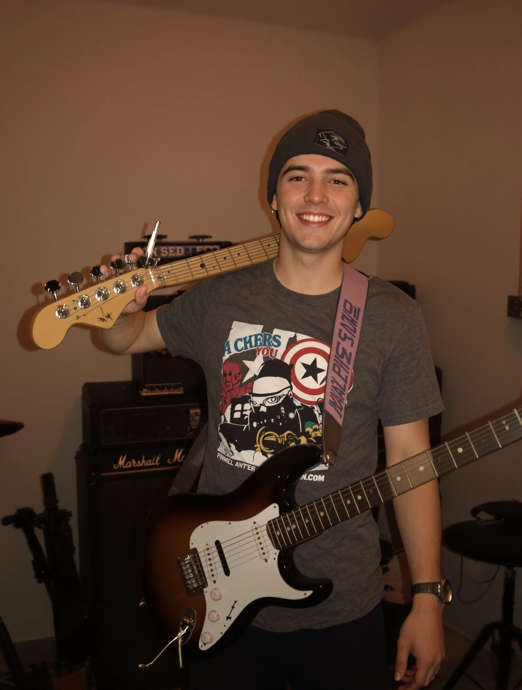 Young man, 18, with his electric guitar slung over his shoulder in a garage band setup, amp behind him, graphic tee and beanie, playful grin, warm tungsten light.