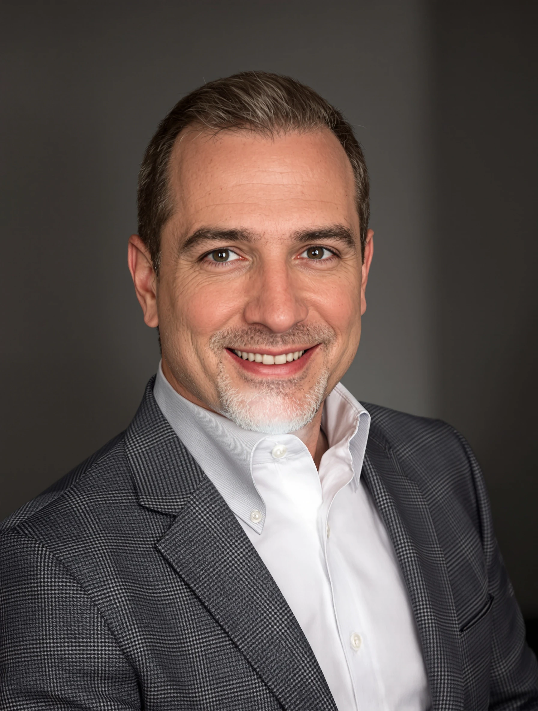 Corporate headshot of a man in a charcoal suit with a white shirt and no tie, soft studio light, polished professional expression.