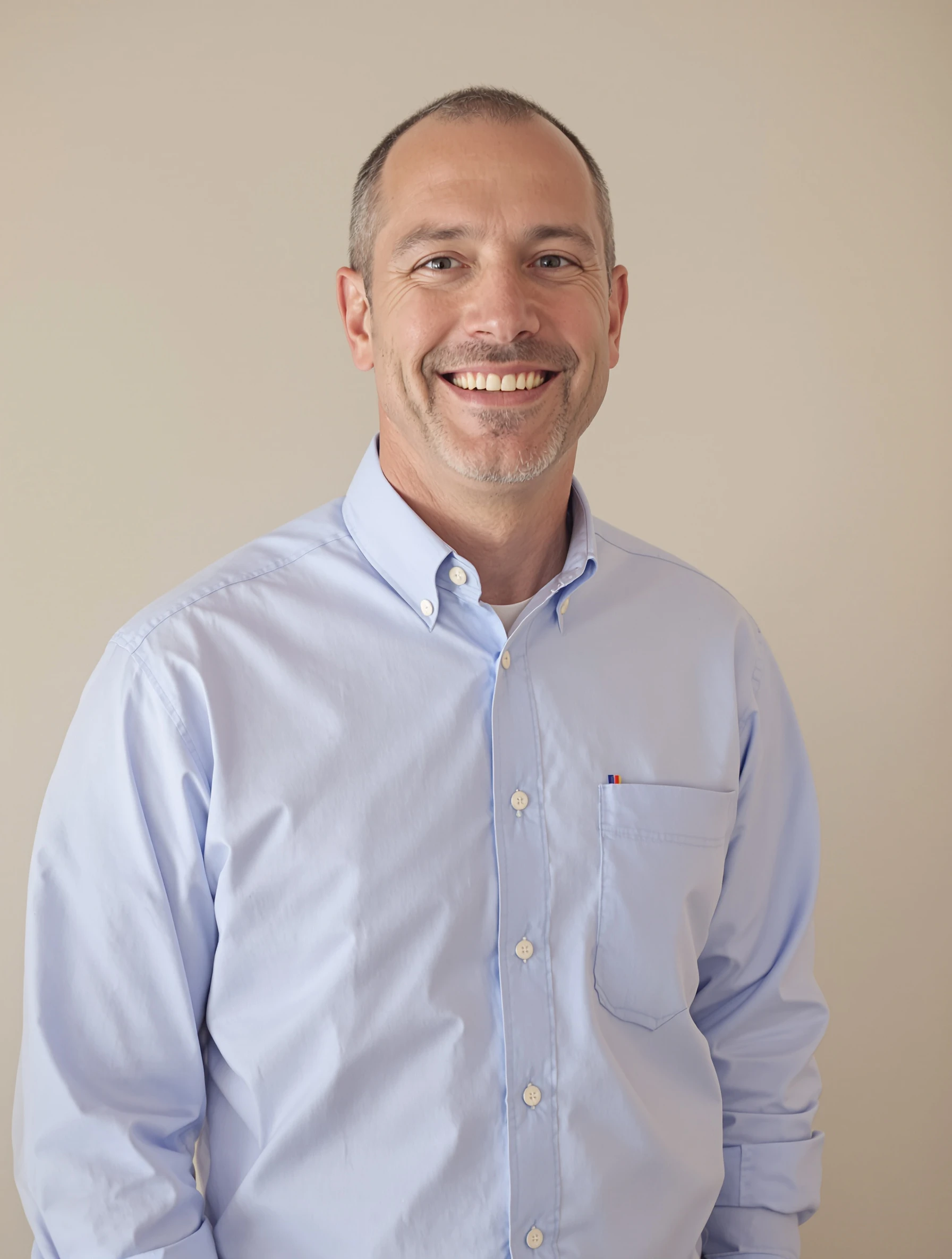 Headshot of a man in a light blue oxford button-down shirt with the top button open, neutral backdrop, friendly approachable smile.