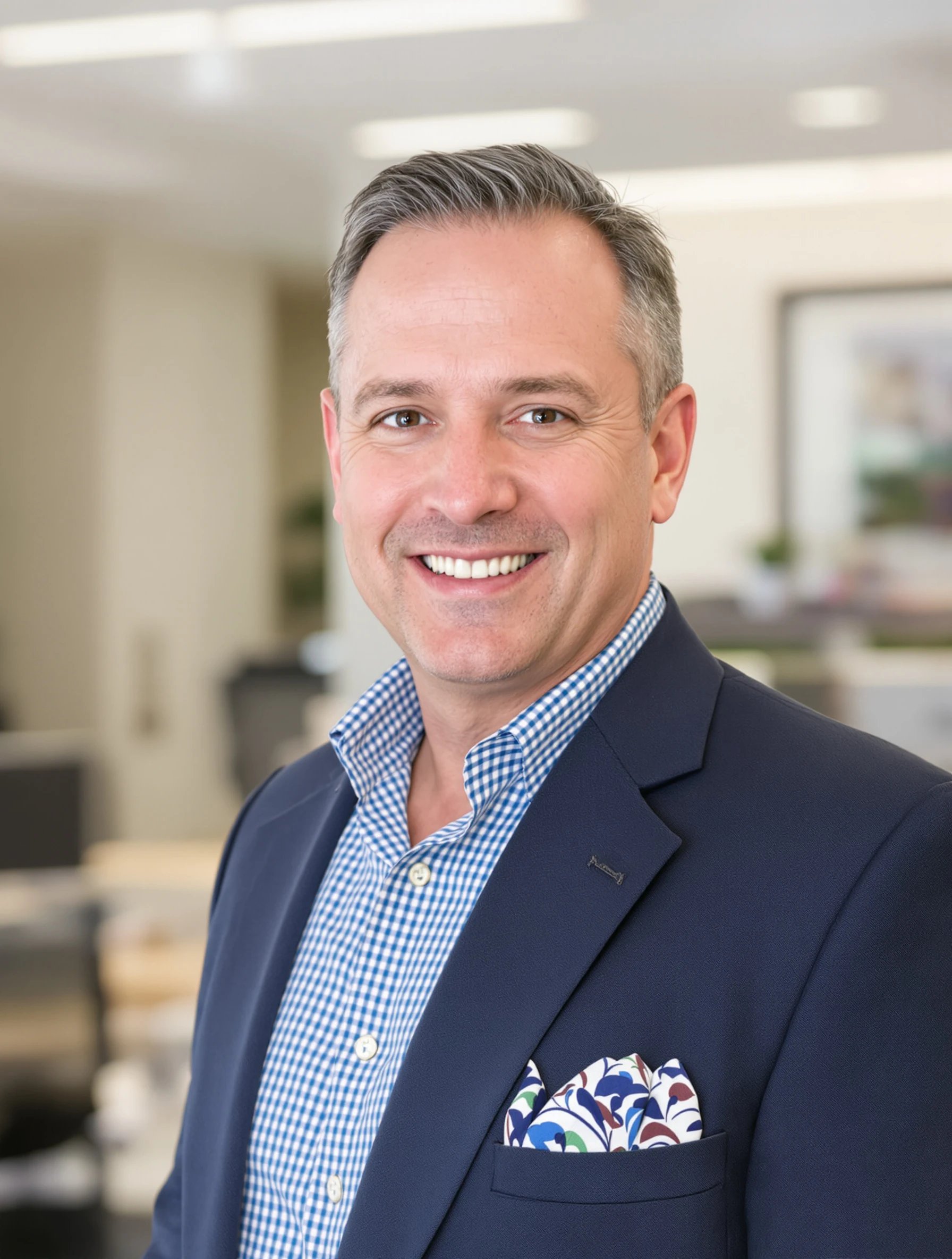 Real estate headshot of a man in a navy blazer with a patterned pocket square and open-collar shirt, bright office backdrop, welcoming smile.