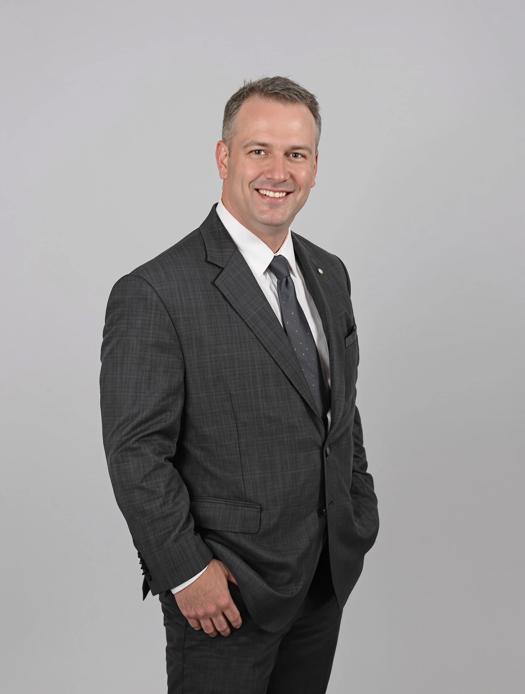 Professional headshot of a man in his 30s, body angled 45 degrees with his face turned toward the lens, charcoal suit with no tie, light grey studio backdrop, confident half smile.