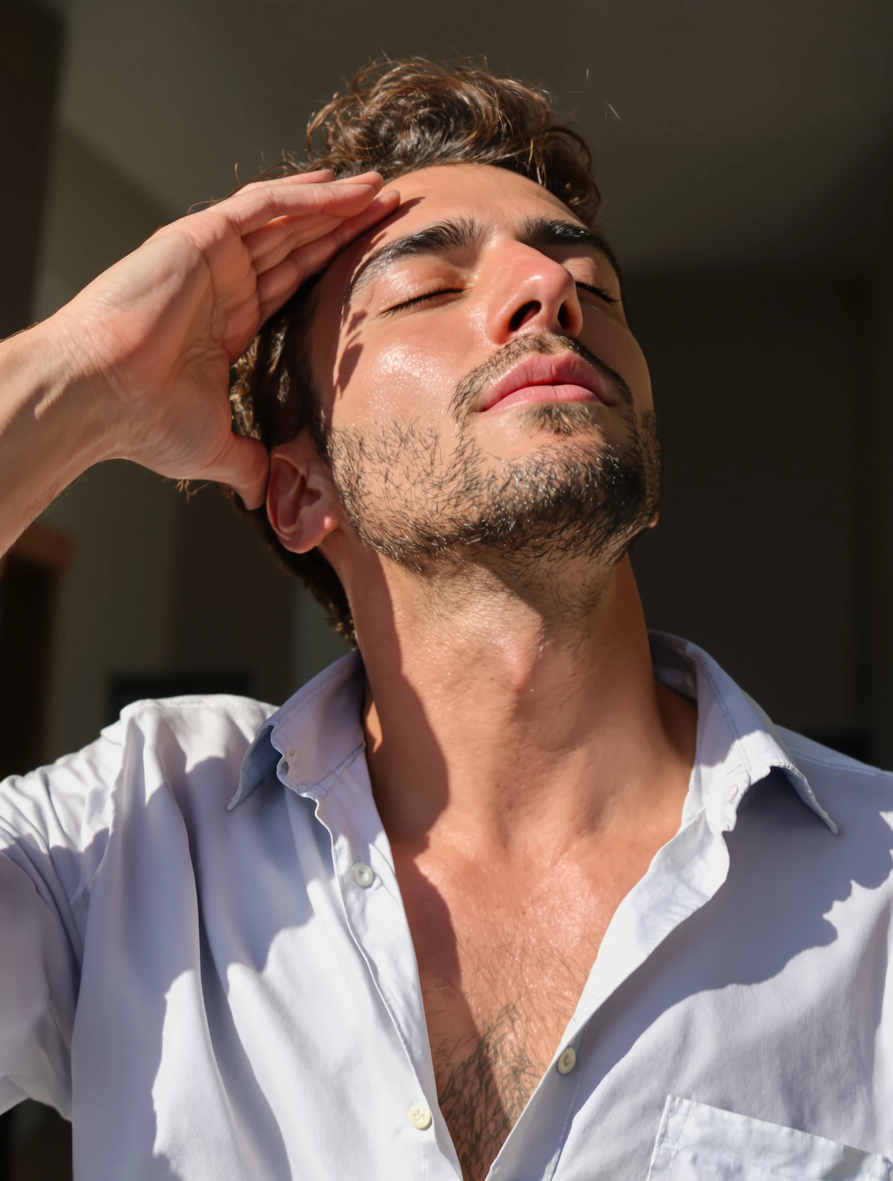Man running a hand through his hair with his eyes closed and chin lifted, sun on his face, open collar shirt, candid relaxed pose.