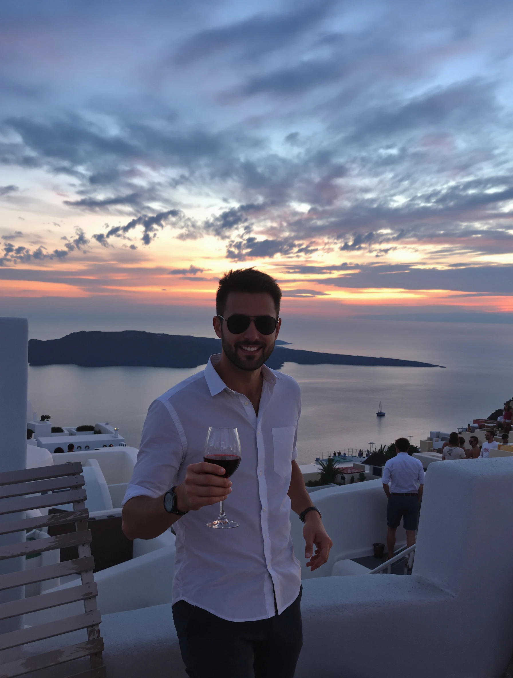 Man enjoying Santorini sunset with wine on caldera-view terrace. Smart casual vacation wear, romantic colors in sky, Greek island relaxation.