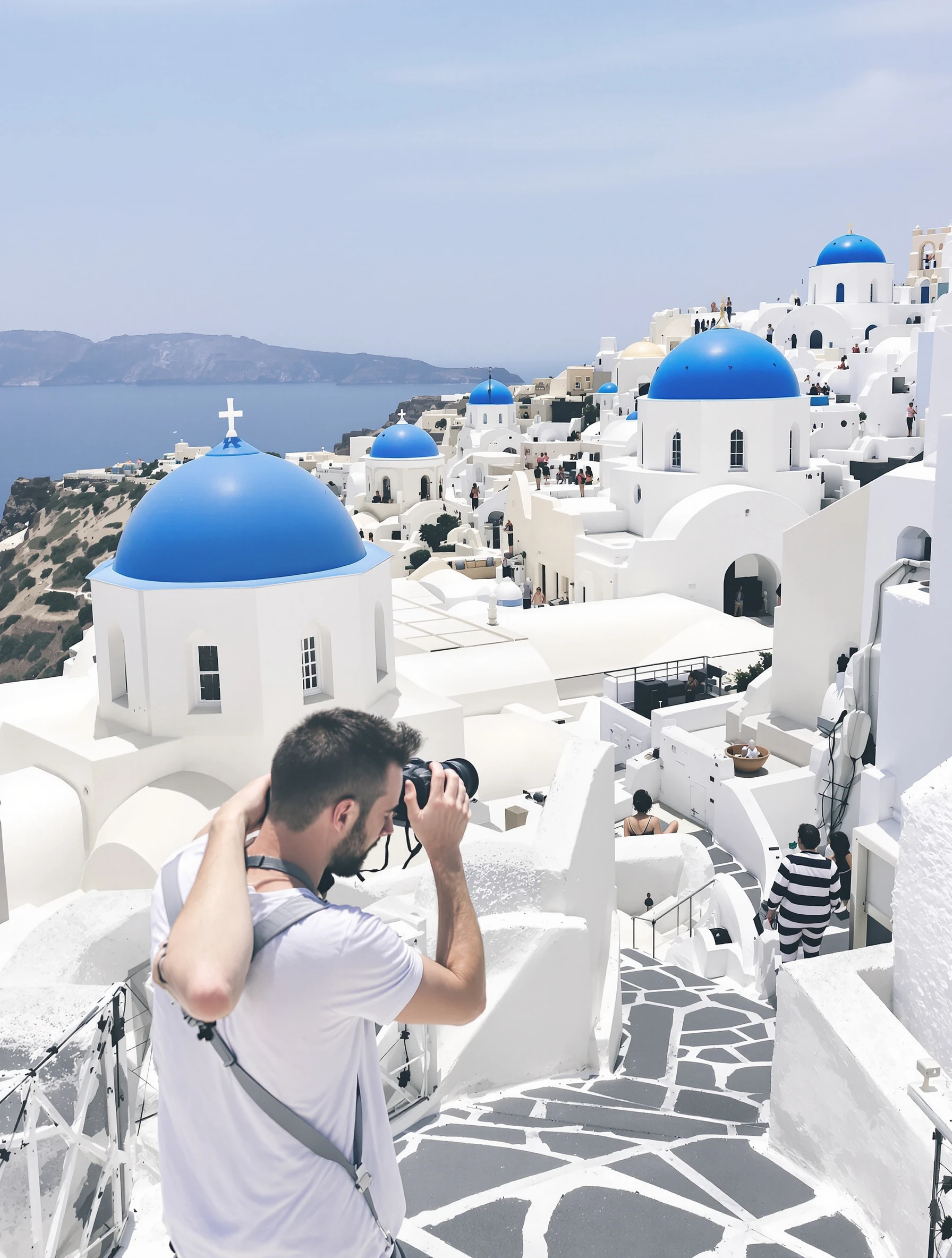 Man photographing iconic Santorini blue domes and white architecture. Casual travel photographer style, camera in hand, chasing the perfect shot.